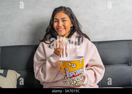 Glückliche junge Frau, die auf dem Sofa sitzt und Popcorn aus dem Eimer isst, während sie zu Hause einen Film ansieht Stockfoto