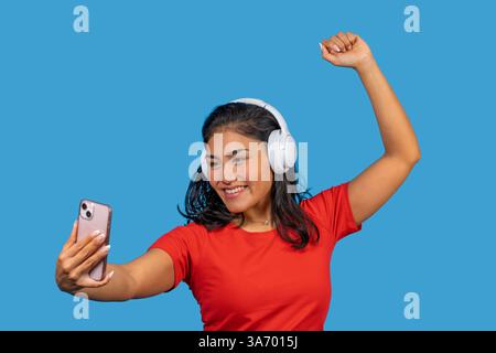 Glückliche lateinfrau, die Musik über Kopfhörer hört, ein Selfie macht und auf blauem Hintergrund tanzt Stockfoto