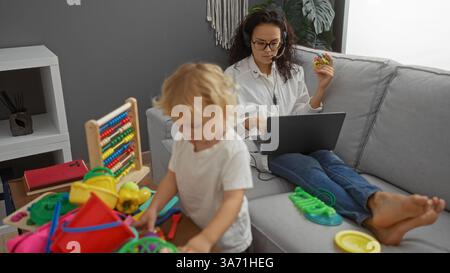Frau, die zu Hause an einem Laptop arbeitet, während der Sohn im Wohnzimmer mit Spielzeug spielt und einen Familienmoment in einem entspannten ap mit einer Frau beim Multitasking zeigt Stockfoto