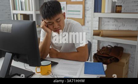 Junger Mann, der in einem Heimbüro sitzt und seinen Kopf auf der Hand vor einem Computer, umgeben von Dokumenten und Büchern, auf einem weißen Schreibtisch Stockfoto
