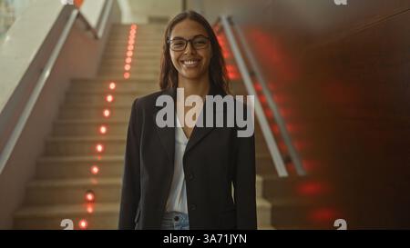 Junge hispanische Frau, die drinnen in der Nähe einer Treppe in einem Lobbykorridor steht Stockfoto