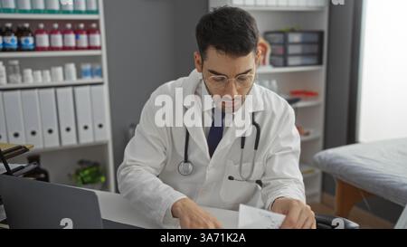 Junger Mann in einer Klinik, der Papierkram überprüft, einen weißen Mantel und Stethoskop trägt, was eine professionelle medizinische Umgebung im Innenraum vorschlägt. Stockfoto