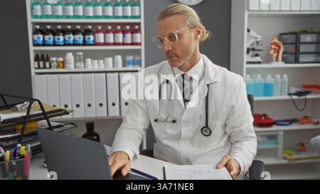 kaukasier mittleren Alters in einem Klinikzimmer, trägt ein Stethoskop, überprüft Dokumente mit einem fokussierten Ausdruck an seinem Schreibtisch, umgeben von medizinischem Su Stockfoto