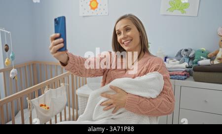 Frau lächelt, während sie Ihr Baby in einem gemütlichen Schlafzimmer hält, um Selfie zwischen farbenfrohen Dekorationen zu machen, die eine warme, fröhliche Atmosphäre im Innenbereich schaffen. Stockfoto