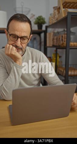 Ein reifer Mann arbeitet nachdenklich am Laptop am Schreibtisch in einer modernen Arbeitsumgebung mit sichtbaren Regalen und Dekor Stockfoto