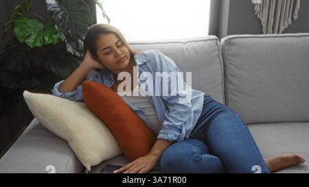 Junge Frau, die sich auf einem Sofa in einem gemütlichen Wohnzimmer mit Pflanzen und Kissen entspannt und eine ruhige Innenumgebung zu Hause darstellt Stockfoto