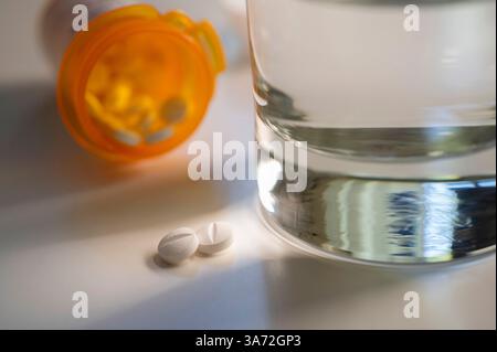 Zwei Pillen, die aus der Pillenflasche und einem Glas Wasser auslaufen Stockfoto