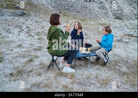 Familie genießt ein Picknick im Freien auf einem Sandfeld, sitzt auf kleinen Campinghockern um einen Tisch, trinkt aus Pappbechern und isst Snacks in einem Stockfoto