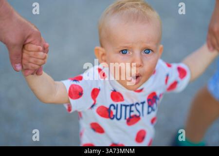 Porträt eines einjährigen Jungen, der in die Kamera blickt, blaue Augen, europäisches Aussehen mit offenem Mund, und er wird von seinen Armen an den Seiten gehalten Stockfoto