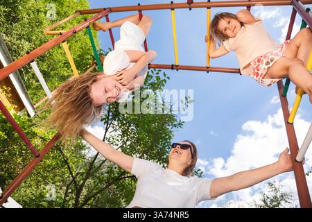 Eine fröhliche Mutter und ihre beiden Töchter spielen in einem bunten Dschungel-Fitnessstudio im Hinterhof, wobei ein Mädchen kopfüber hängt und alle lächeln Stockfoto