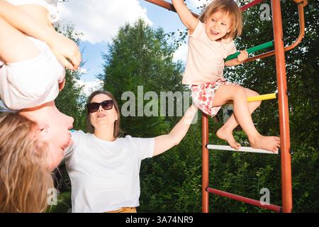 Eine lächelnde Mutter interagiert mit ihren beiden Töchtern, während sie klettern und in einem farbenfrohen Dschungelstudio in einem sonnigen Hinterhof hängen und einen lebendigen Moment der Sum genießen Stockfoto