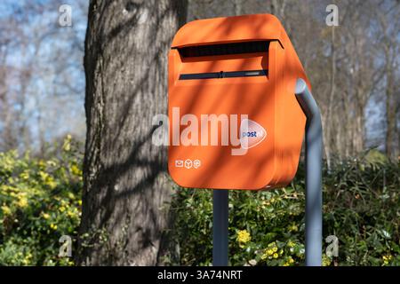 Typischer holländischer orangefarbener Briefkasten mit PostNL-Logo, der sich im Frühjahr in einer Straße entlang eines Parks mit Bäumen und Sonne befindet Stockfoto