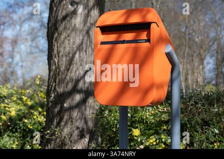 Orangefarbener Briefkasten draußen in einer Straße entlang eines Parks mit Bäumen im frühen Frühling mit Sonne. Schatten von Bäumen und Sträuchern. Keine Zeichen Stockfoto