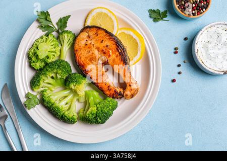 Gegrilltes Lachsfischsteak mit gedämpftem Brokkoli-Gemüse und Joghurtsauce auf blauem Hintergrund, Blick von oben. Gesunde ausgewogene Ernährung Konzept, Keto Diät. Stockfoto