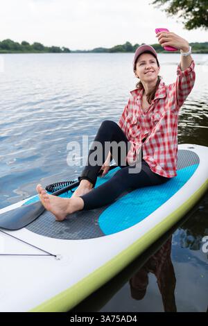 Eine Frau in einem roten karierten Hemd und Leggings, die auf einem Paddleboard sitzt und lächelt, während sie ein Selfie auf einem ruhigen See macht. Stockfoto