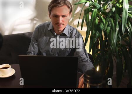 Mann in Hemd in Geschäftskleidung arbeitet am Computer, Home Office, gemütliche Atmosphäre, Palme bequemes Sofa. Stockfoto