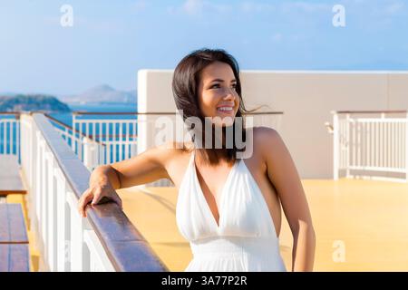Eine hübsche junge brünette Frau in einem weißen Sonnenkleid lächelnd auf dem Oberdeck eines Kreuzfahrtschiffs im Mittelmeer. Stockfoto