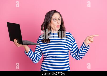 Eine ältere Frau, die mit verspielten Gesten einen Laptop hält, steht vor einem leuchtend rosa Hintergrund Stockfoto