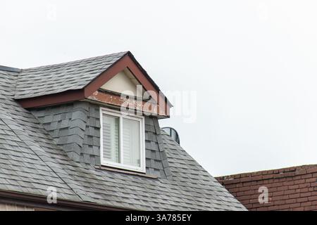 Detaillierte Ansicht eines Dachdormers mit grauen Schindeln, einem weiß gerahmten Fenster und abblätternder Farbe auf der Verkleidung. Stockfoto