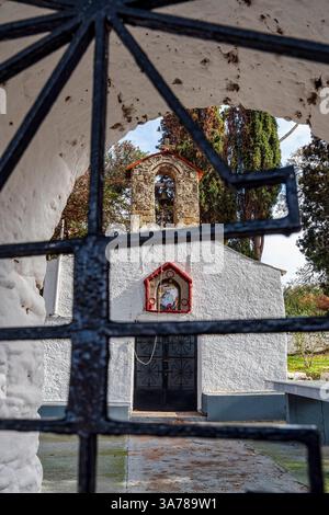 Blick auf eine traditionelle, weiß getünchte griechische Kapelle durch ein dekoratives Eisentor mit Glockenturm und religiöser Ikonographie. Stockfoto