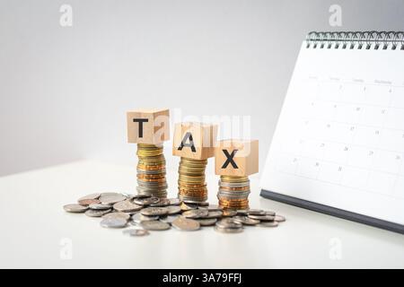 Steuerwort auf Holzblöcken auf Münzen, die auf einem Tisch liegen, mit Kalender im Hintergrund. Stockfoto