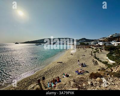 Sonnenanbeter genießen die Sonne am späten Nachmittag am Strand Platis Gialos auf der Insel Mykonos, Griechenland, mit Branco Beach Club in der Ferne. Stockfoto