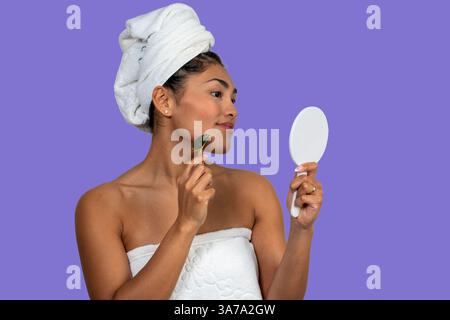 Eine junge Frau, die ein Badetuch trägt und einen Gua-Sha-Stein im Gesicht trägt und in einen kleinen Handspiegel blickt Stockfoto