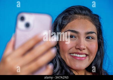 Fröhliche hispanische Frau, die ein Selfie mit Handy vor blauem Hintergrund macht Stockfoto