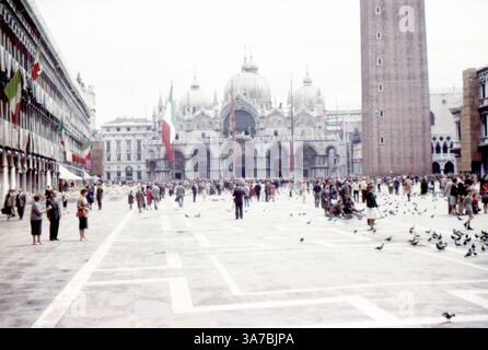 Markusplatz (Piazza San Marco) in Venedig, Italien, fotografiert im Juli 1965. Diese lebhafte Szene zeigt die ikonische Fassade des Markusdoms mit seinen reich verzierten Kuppeln, den majestätischen Campanile di San Marco und die Besuchermassen, die sich unter Tauben auf dem Platz mischen. Italienische Flaggen hängen an den nahe gelegenen Gebäuden und verleihen dem Nachkriegszauber einen festlichen Charme. Das Bild, das auf Diafilm aufgenommen wurde, spiegelt die lebendige Atmosphäre und klassische Architektur des Venedig der 1960er Jahre wider und bietet einen nostalgischen Einblick in die europäische Reise der Mitte des Jahrhunderts. Stockfoto