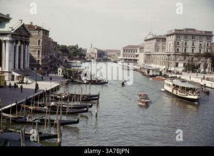 Ein malerischer Blick auf den Canal Grande in Venedig, aufgenommen im Jahr 1965, mit historischen Palästen, Gondeln und Wassertaxis der Stadt. Der Kanal, gesäumt von eleganter venezianischer Architektur, schlängelt sich durch das Herz der Stadt und dient als Hauptwasserstraße. Gondoliere navigieren geschickt durch den Kanal, während Vaporettos (öffentliche Wasserbusse) Passagiere durch diese zeitlose Landschaft transportieren. Stockfoto