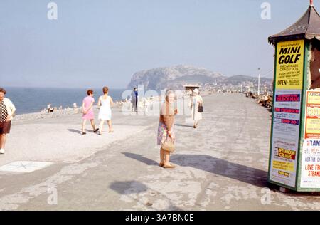 Urlauber schlendern an einem sonnigen Tag in den 1960er Jahren entlang der Promenade in Craig-y-Don in Llandudno, Wales Diese lebendige Küstenszene, die auf 35-mm-Farbdiafilm aufgenommen wurde, umfasst Strandbesucher, klassische Kleidung und Vintage-Schilder, die Minigolf und Küstenrundfahrten Werbung machen. Stockfoto