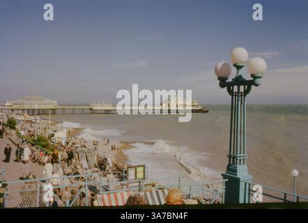 Ein lebhaftes Foto aus den 1960er Jahren des Eastbourne Pier, aufgenommen auf originalem 35-mm-Farbdiafilm. Das Bild zeigt Strandbesucher, die sich in Liegestühlen entlang der Promenade entspannen, während sich der historische Pier unter klarem blauen Himmel bis ins Meer erstreckt Stockfoto