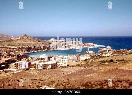 Ein atemberaubender Blick über die Marsalforn Bay auf der Insel Gozo, Malta, 1960er Jahre. Dieses Vintage-Foto fängt den ruhigen Charme des Küstendorfes vor der großangelegten Entwicklung mit seinen traditionellen Kalksteinhäusern mit Flachdach, einem kleinen Kirchturm und dem tiefen Blau des Mittelmeers bis zum Horizont ein. Im Hintergrund tragen die geschwungene Küste von Mgarr ix-Xini und die umliegende ländliche Landschaft zur zeitlosen, ruhigen Atmosphäre der Insel bei. Stockfoto