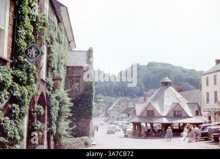 Die Dunster High Street in Somerset ist eine lebendige Szene aus den 1960er-Jahren mit dem legendären achteckigen Garnmarkt im Zentrum. Einheimische und Besucher treffen sich in der Nähe von Oldtimern, während Evy die Mauern des historischen Luttrell Arms Hotel auf der linken Seite erklimmt. Stockfoto