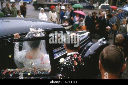 Ein frisch verheiratetes Paar steigt in ein Auto, während Hochzeitsgäste sie vor dem Veranstaltungsort mit Konfetti übergießen. Fotografiert im Juli 1963 auf 35-mm-Diafilm. Stockfoto