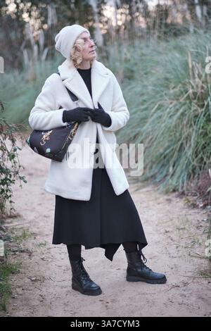 Modische Frau mit weißem Mantel, schwarzen Handschuhen und Stiefeln, die einen friedlichen Spaziergang in der Natur genießt Stockfoto