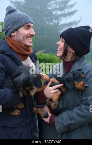 Junge Paare lachen und halten ihre beiden liebenswerten Hunde in einem nebeligen Park an einem kalten Wintertag Stockfoto