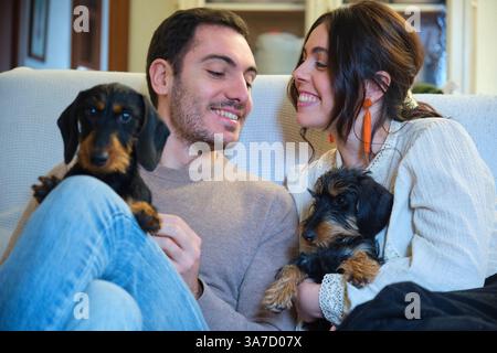Junges, liebevolles Paar, das die Zeit zusammen auf dem Sofa mit ihren zwei bezaubernden dackelhaarigen Hunden genießt Stockfoto