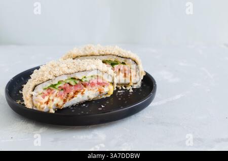 Sushi-Burger mit Thunfisch, Gurke, Zwiebeln, Käse und Reis in Nori mit knuspriger Kruste auf einem schwarzen Teller auf grauem Hintergrund. Kreatives Gericht. Hybridfutter c Stockfoto