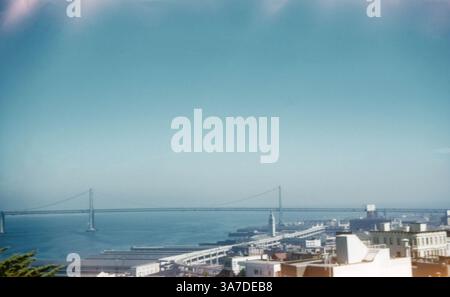 Ein Farbdiafoto aus den 1960er Jahren bietet einen klaren Blick auf die San Francisco–Oakland Bay Bridge, die sich über die Bucht erstreckt. Von einem erhöhten Aussichtspunkt in San Francisco aus aufgenommen, zeigt das Bild die ursprüngliche, doppelstöckige östliche Brücke, darunter der Embarcadero- und Ferry Building-Uhrenturm. Stockfoto