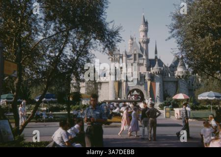 Foto aus den 1960er Jahren des berühmten Dornröschenschlosses in Disneyland, Kalifornien, vom Eingang zu Fantasyland aus gesehen. Besucher schlendern entlang der Main Street in Vintage-Kleidung, während ein klassischer gelber Omnibus die Gäste zu den Schlosstoren bringt. Stockfoto