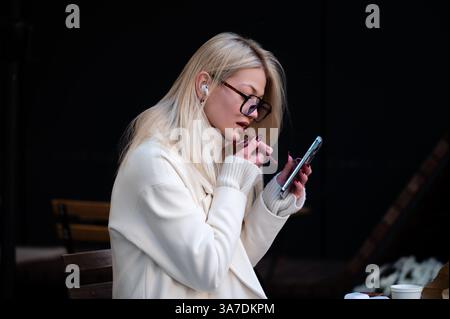 Die langhaarige Frau in einem schicken weißen Mantel trägt Lippenstift auf, während sie auf ihr Smartphone blickt und in einen Moment der Selbstpflege und digitalen Verbindung eingetaucht ist Stockfoto