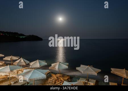 Ein friedlicher, mondbeleuchteter Strand mit leeren Liegestühlen und Strohschirmen unter einem strahlenden Nachthimmel. Das Mondlicht reflektiert vom ruhigen Meer und schafft einen Ser Stockfoto