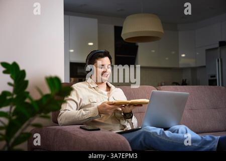 Mann, der auf dem Sofa sitzt und Pizza isst, während er am Laptop arbeitet Stockfoto