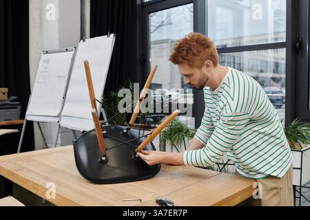 Ein junger Mann konzentriert sich darauf, einen Stuhl in einem modernen Arbeitsbereich zusammenzustellen, während er umzieht. Stockfoto