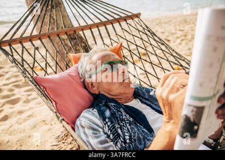 Senior Mann löst Rätsel in der Zeitung, während er sich während des Urlaubs auf einer Hängematte am Strand liegend Stockfoto