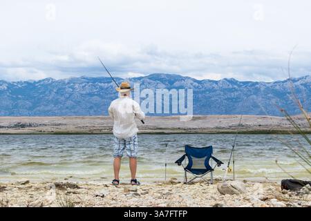Rückansicht eines alten, nicht erkennbaren Fischers, der einen Strohhut trägt und eine Angelstange hält Stockfoto