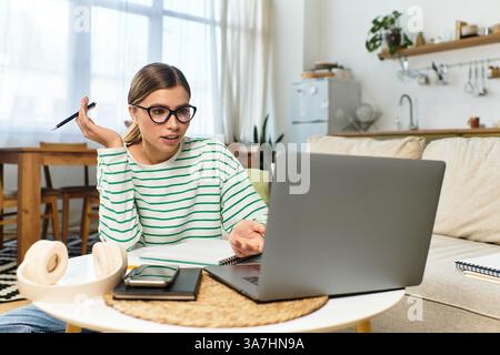 Eine junge, schöne Frau sitzt an ihrem gemütlichen Arbeitsplatz, konzentriert sich auf ihren Laptop, während sie Notizen macht. Stockfoto