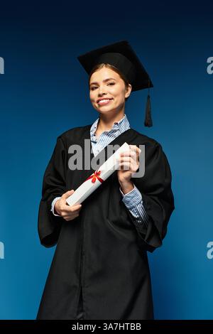 Eine junge Frau hält mit Freude ihr Diplom in ihrem Abschlusskleid und ihrer Mütze, um den Erfolg zu feiern. Stockfoto