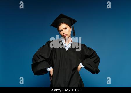 Eine stolze junge Frau in Abschlusskleidung steht selbstbewusst vor einem kühnen blauen Hintergrund. Stockfoto